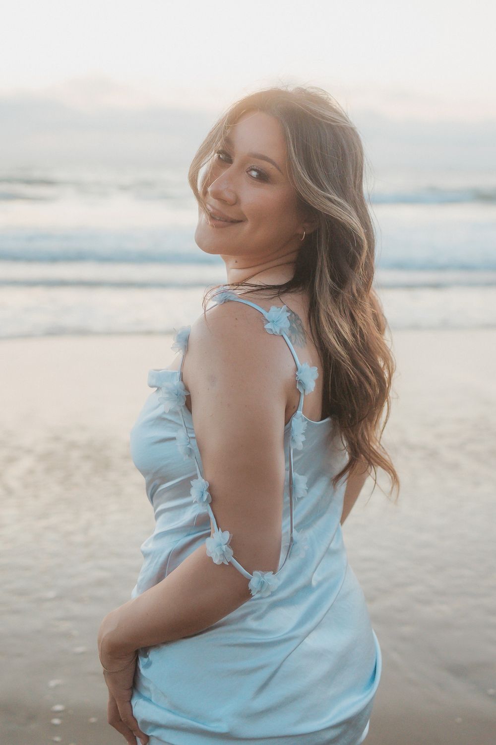 Young woman in light blue ruffled top poses on the beach at sunset against a cloudy sky.
