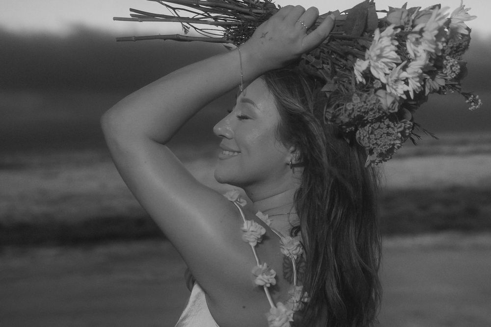 A woman wearing a flower crown smiles peacefully with her hand raised against a moody sky in black and white.