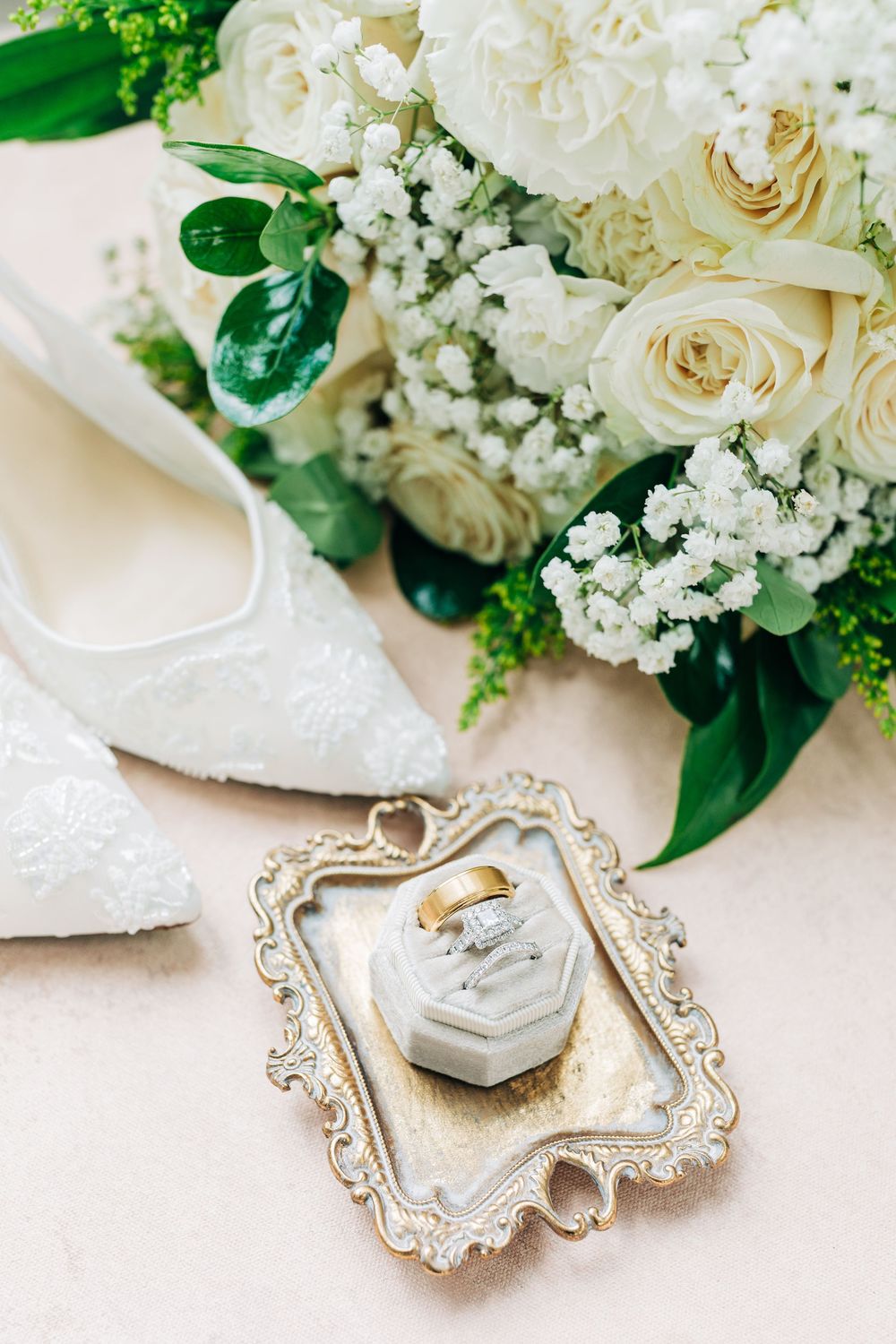 Vintage-style wedding accessories with white floral bouquet, ornate silver frame, and bridal shoes on beige surface.