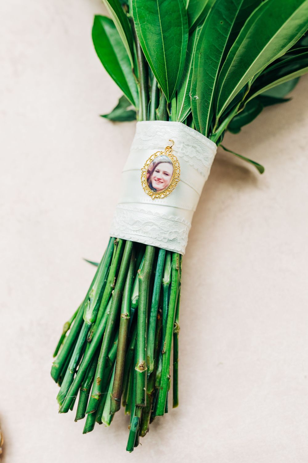 Wedding bouquet stems wrapped in white ribbon and decorated with a vintage-style cameo brooch accent against light background.