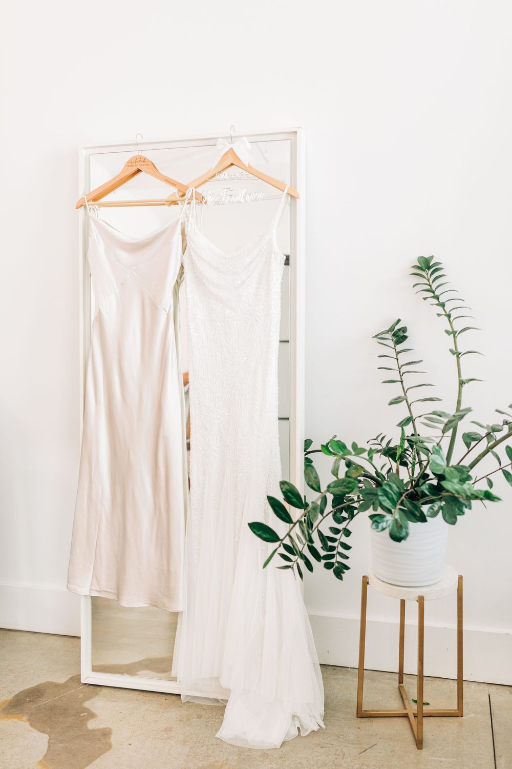 A white wedding dress and slip garments hanging on wooden hangers in a bright minimalist space with green foliage accents.