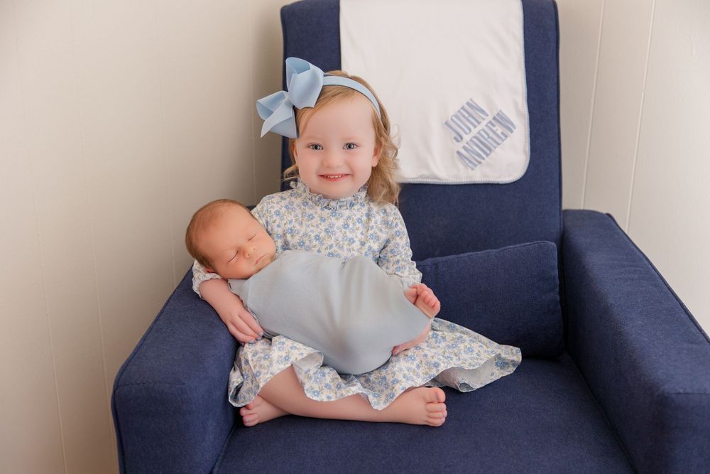 Two babies wearing matching gray floral outfits sit together on a navy blue chair.
