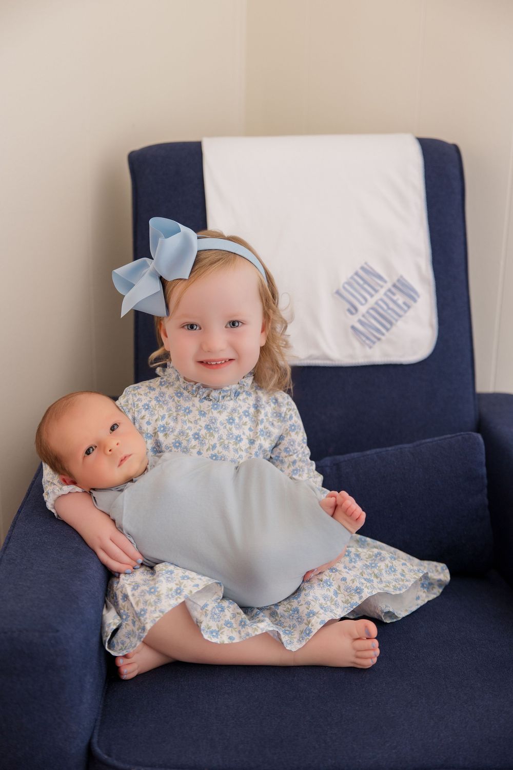 A baby in a blue floral dress lies playfully across a navy blue couch in five sequential photos.