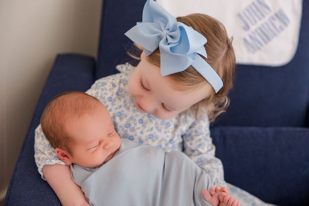 A newborn baby wearing a blue bow sleeps peacefully while resting on a dark blue sofa.