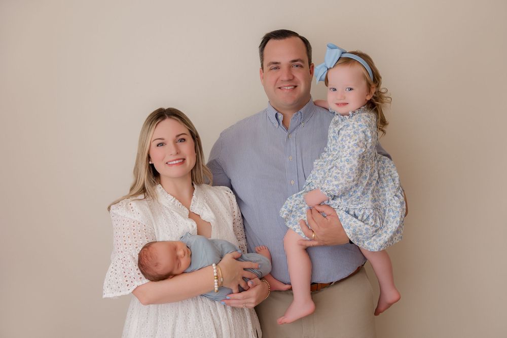 Family of four poses together in front of beige wall wearing coordinated white and blue outfits.