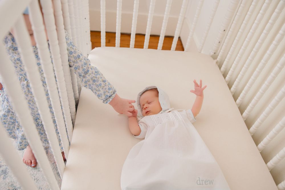 Sleeping baby in white sleepwear stretches inside a wooden crib with grey patterned bedding.
