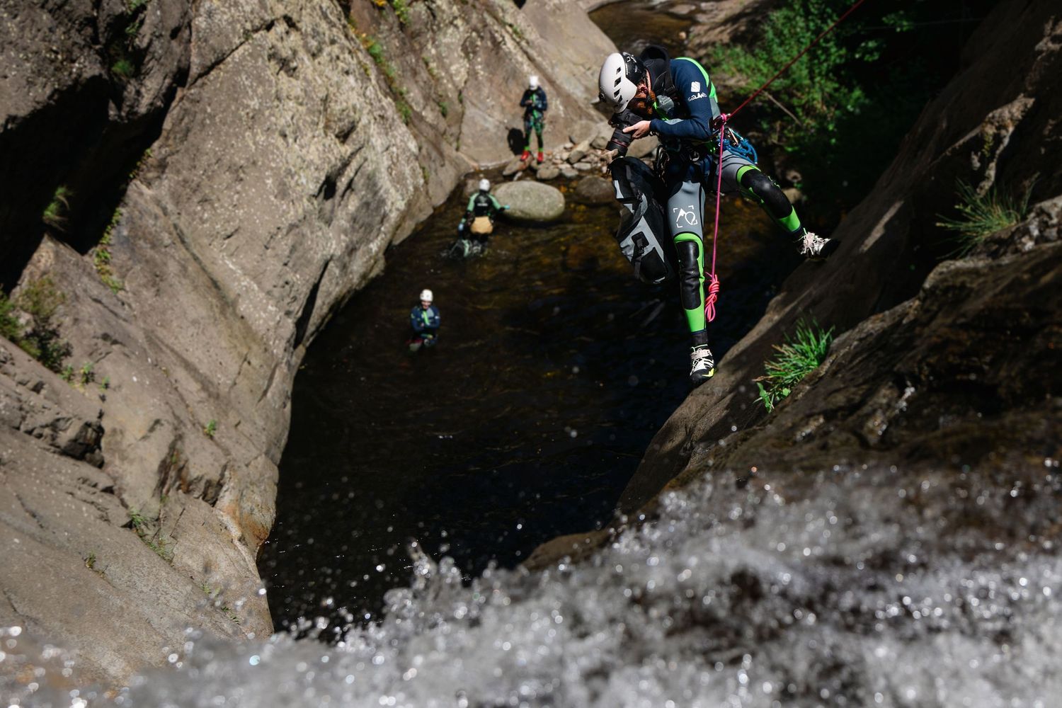 Expedition photographer in a wetsuit on a rope taking photos of a waterfall cascading down rocky cliff with climbers in the background.