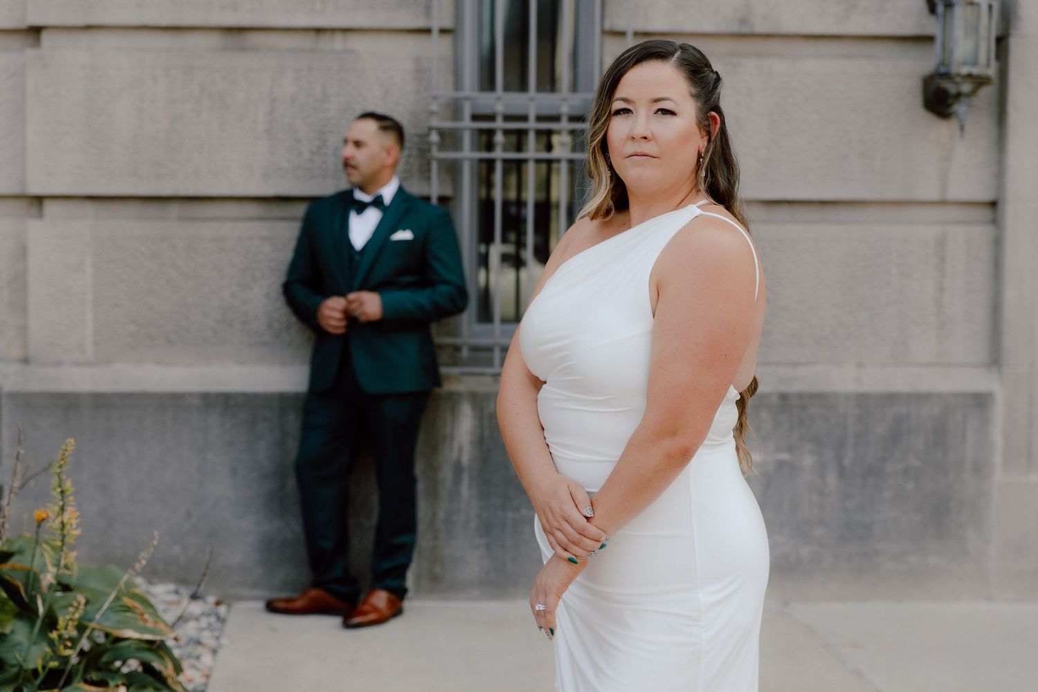 A couple poses together in formal wedding attire in front of a stone building exterior.