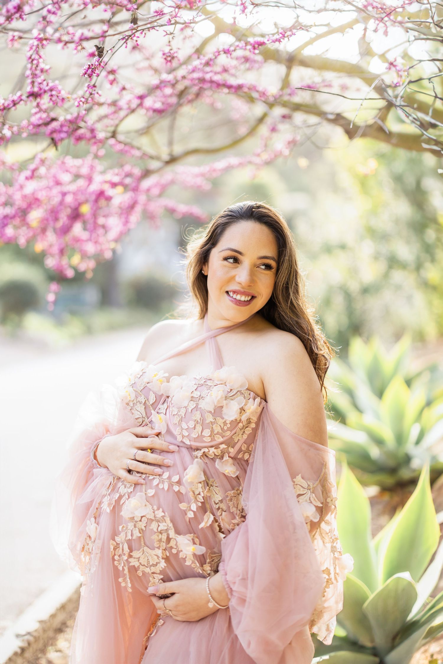 Maternity portrait under blooming cherry blossoms in soft pink lighting.