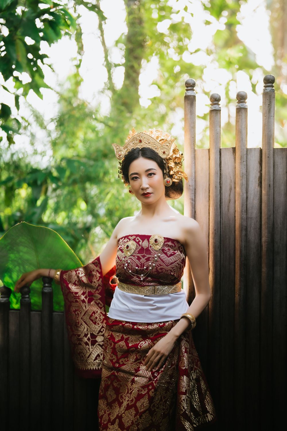 Figure in burgundy traditional dress poses gracefully against a wooden fence with sunlit foliage.