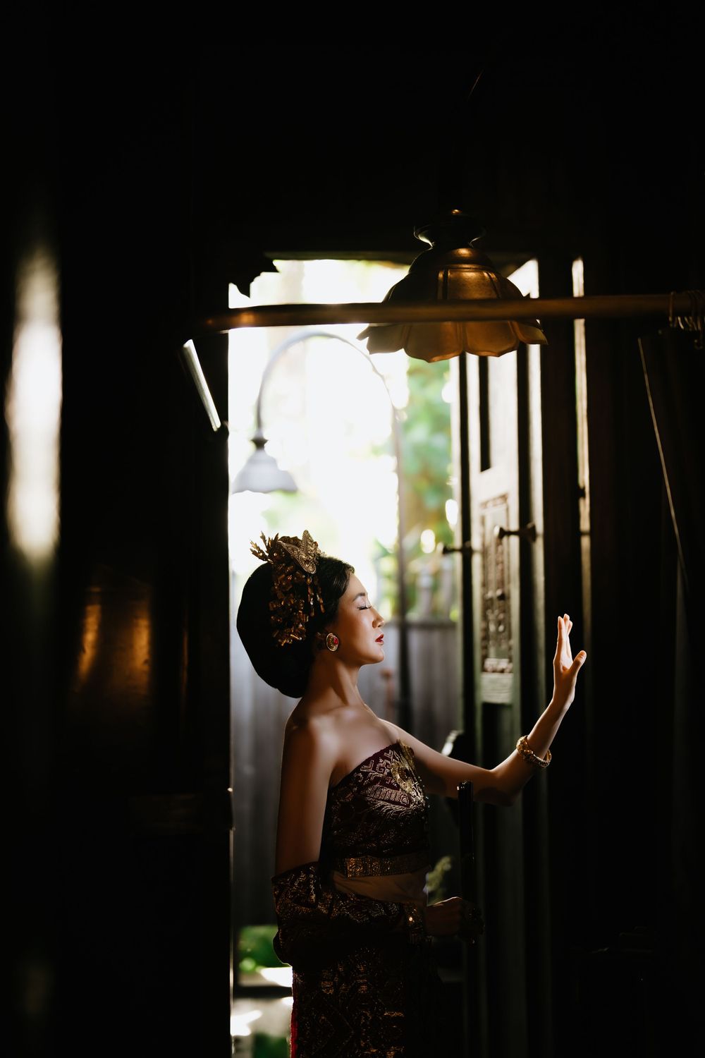 A dramatic portrait in dark lighting shows a figure in traditional dress looking upward near a window.
