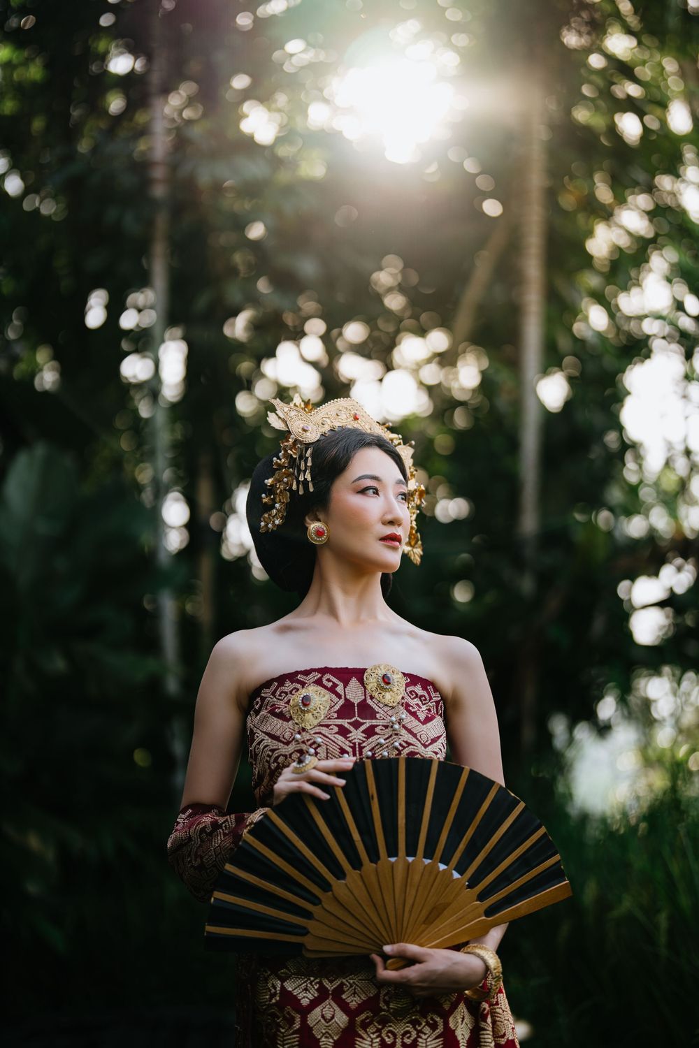 Traditional Asian-inspired photoshoot in forest setting with decorative fan and burgundy dress.