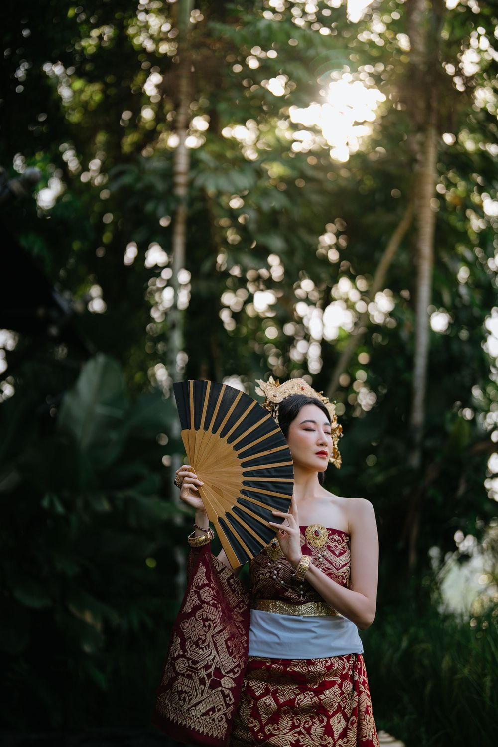 Artistic portrait with folding fan in sunlit forest wearing traditional burgundy attire.