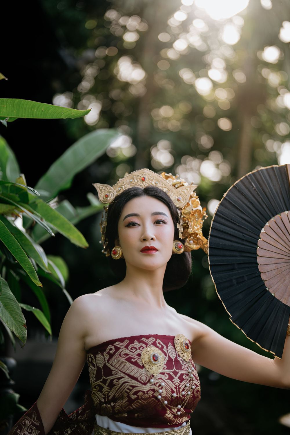 Elegant portrait with decorative fan and ornate headpiece in dappled forest light.