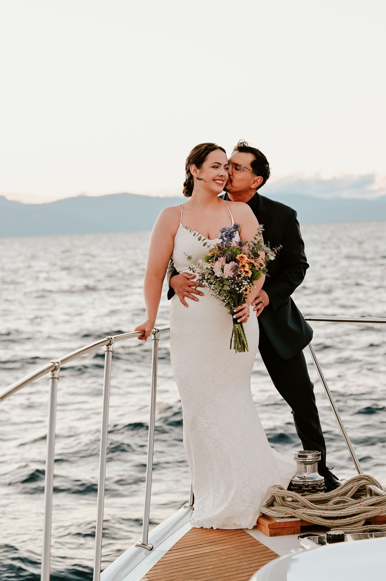 A romantic couple shares intimate moments on a boat deck with mountains and water in the background at sunset.