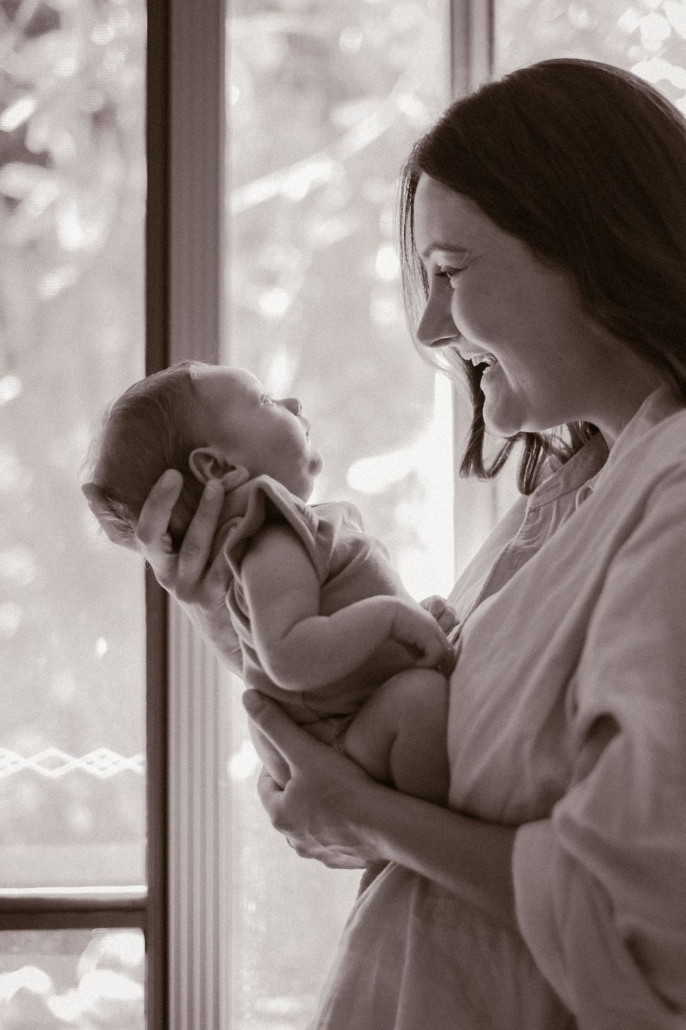 A mother in white holding her newborn baby by a window with soft natural light streaming in.