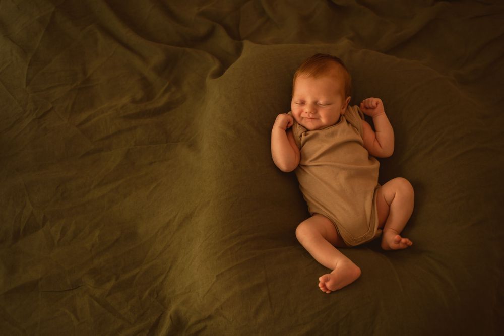 Newborn baby wearing beige onesie lying on dark olive green fabric.