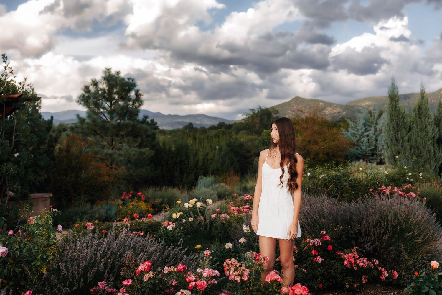 Young woman in white dress stands among colorful blooming garden under dramatic cloudy sky.