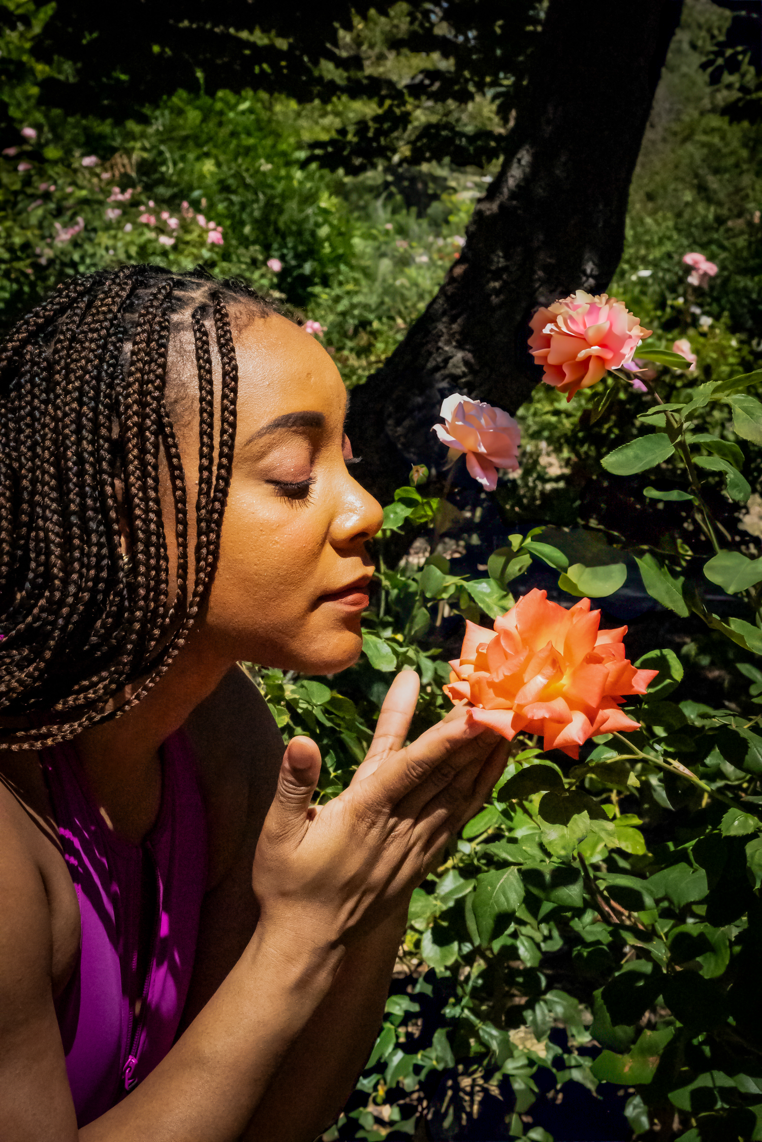 Close-up profile of someone smelling a vibrant coral rose in a garden setting.