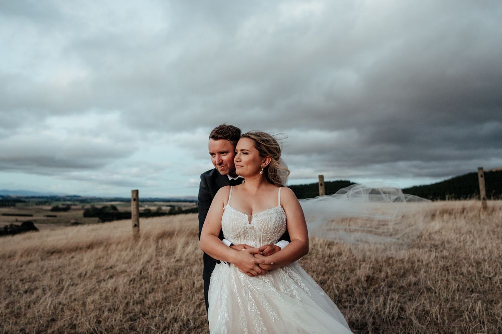 Bride and groom stand together on a windswept golden hill beneath dramatic stormy clouds at Tironui Farm Martinborough by August and Vine