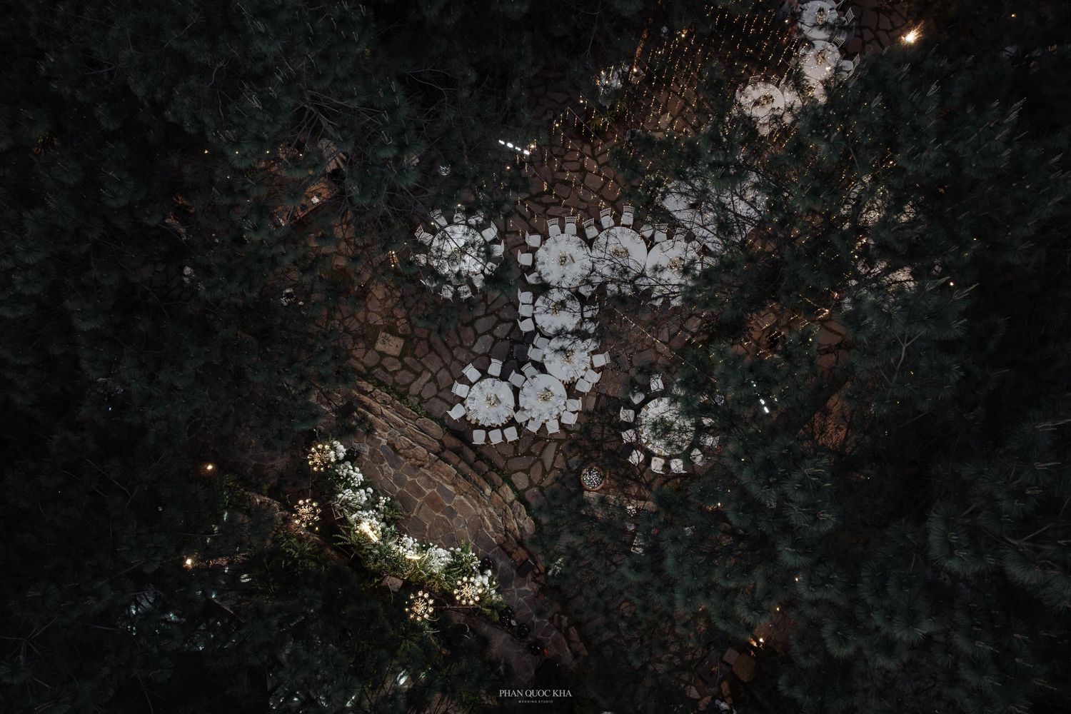 Aerial night view of outdoor wedding reception with round white tables glowing among dark pine forest.