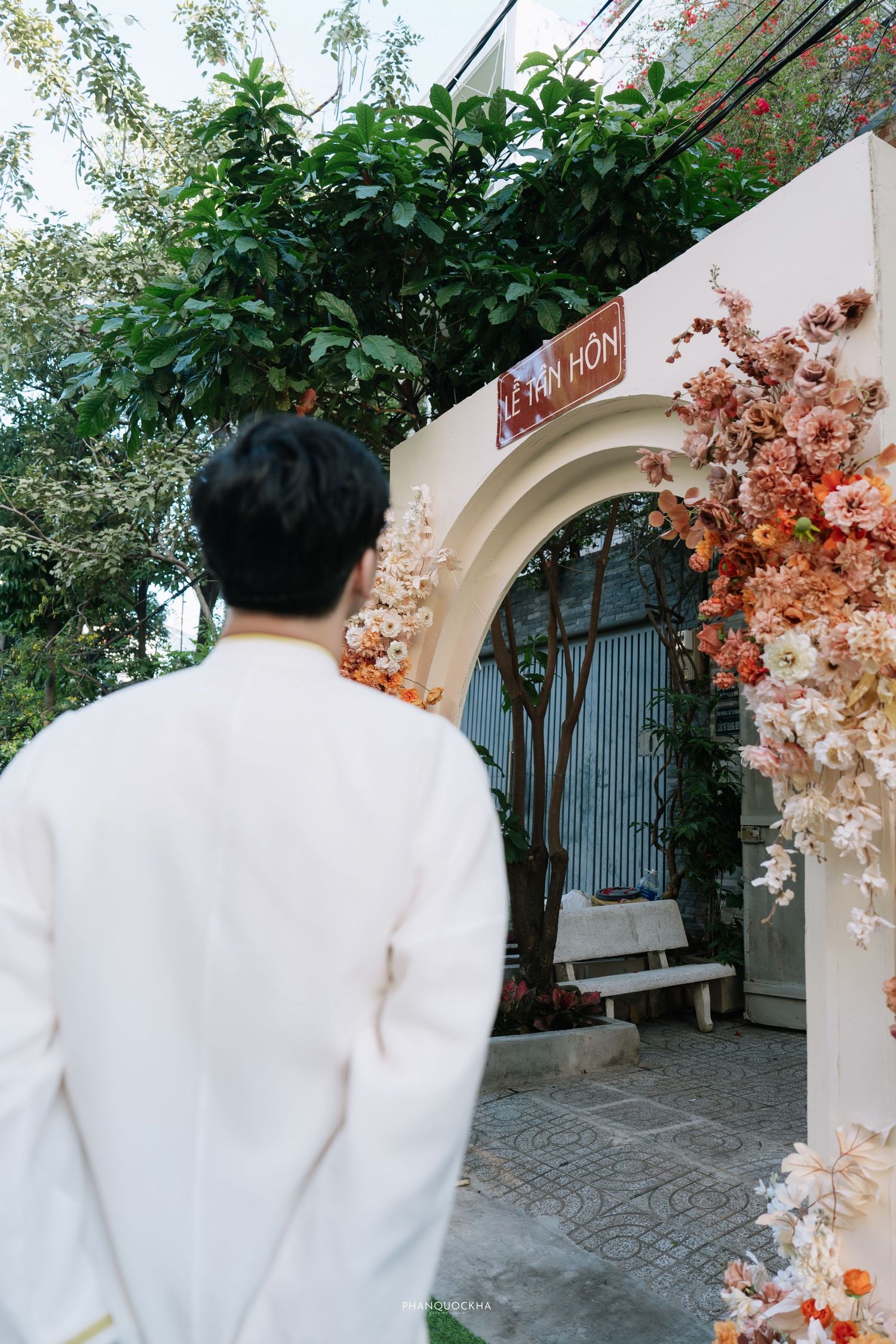 Groom in white ao dai walks toward floral wedding arch decorated with pink and orange flowers.