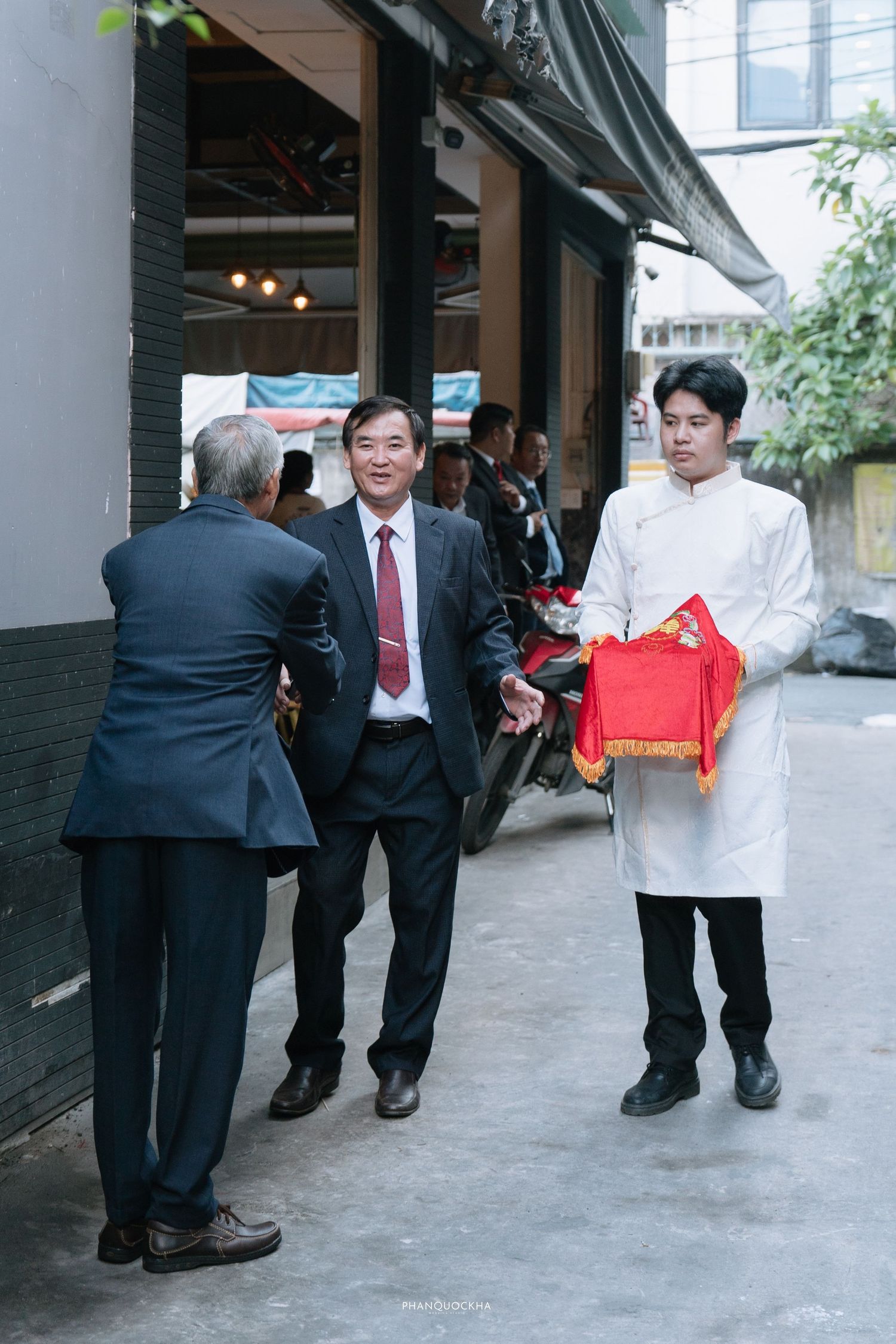 Man in white ao dai holds red ceremonial gift box while speaking with suited guests outdoors.