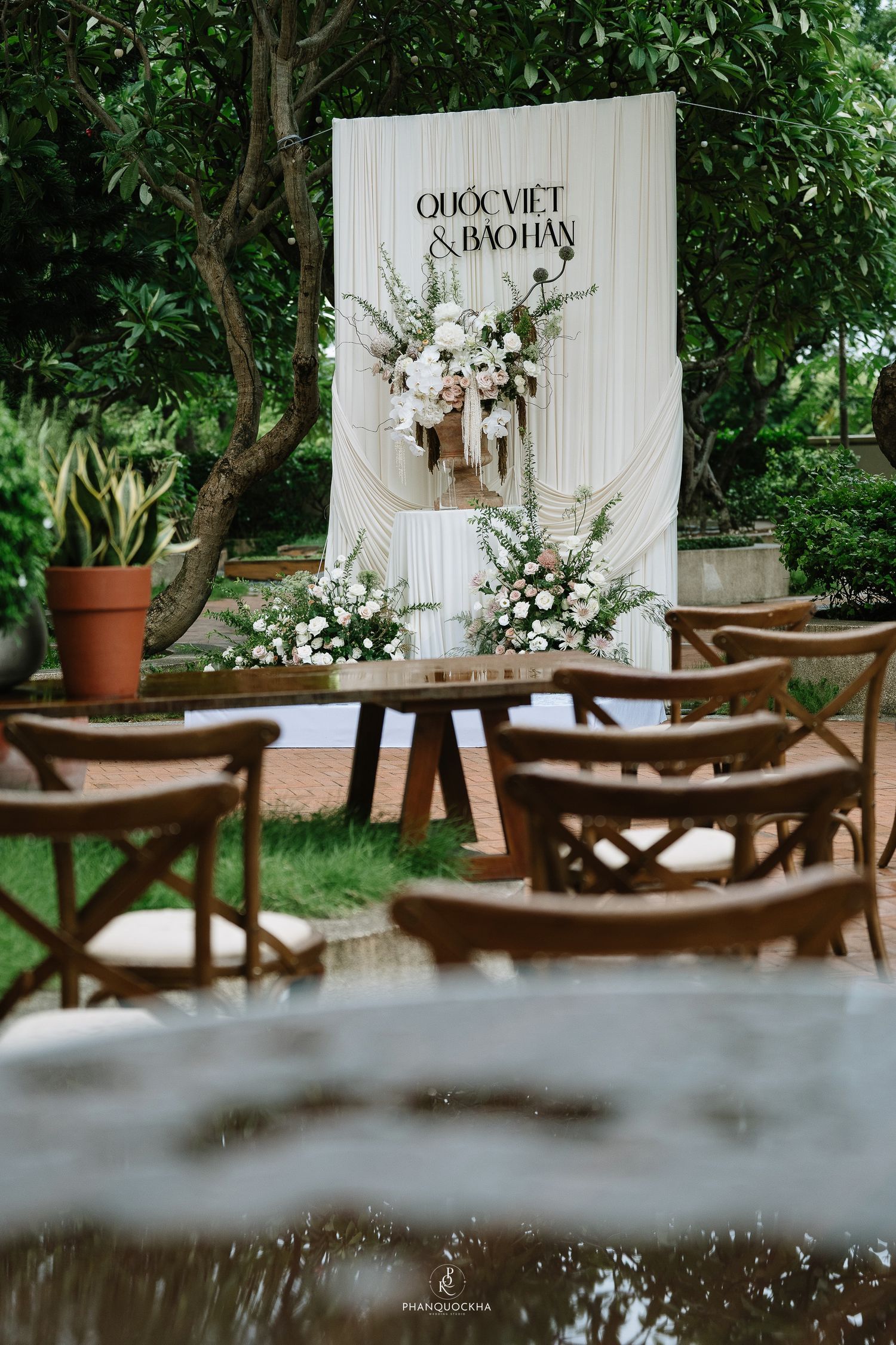 Elegant outdoor wedding backdrop for Quoc Viet and Bao Han with white draping and floral arrangements.