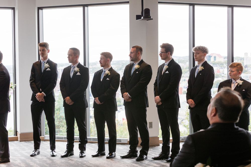 Wedding party members in black suits stand in line against bright windows.