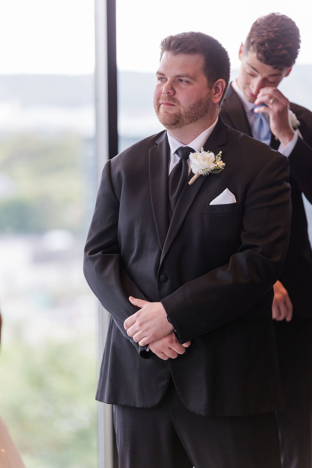 Wedding attendee in formal black suit and boutonniere wipes away tears during emotional moment at ceremony.