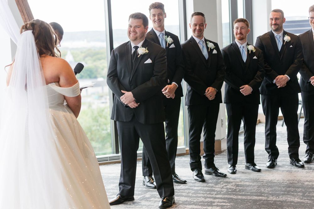 Wedding ceremony participants arranged in formal line with large windows in background.