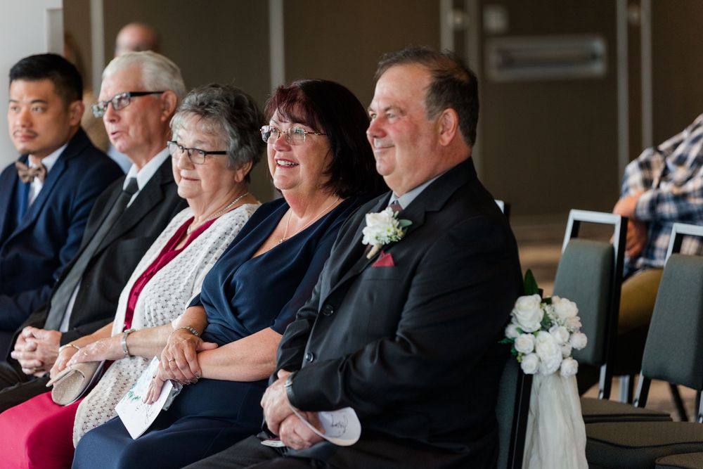 Wedding guests seated in elegant attire observe a ceremony in a bright venue with large windows.
