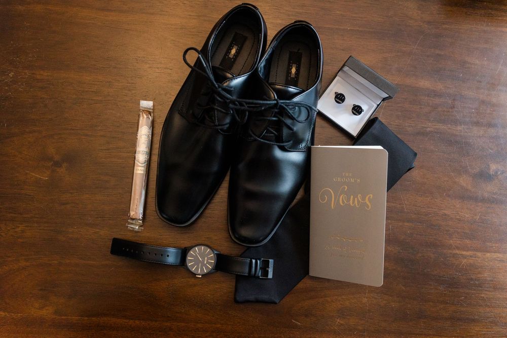 Black leather dress shoes and accessories arranged on dark wooden surface with wallet and watch.
