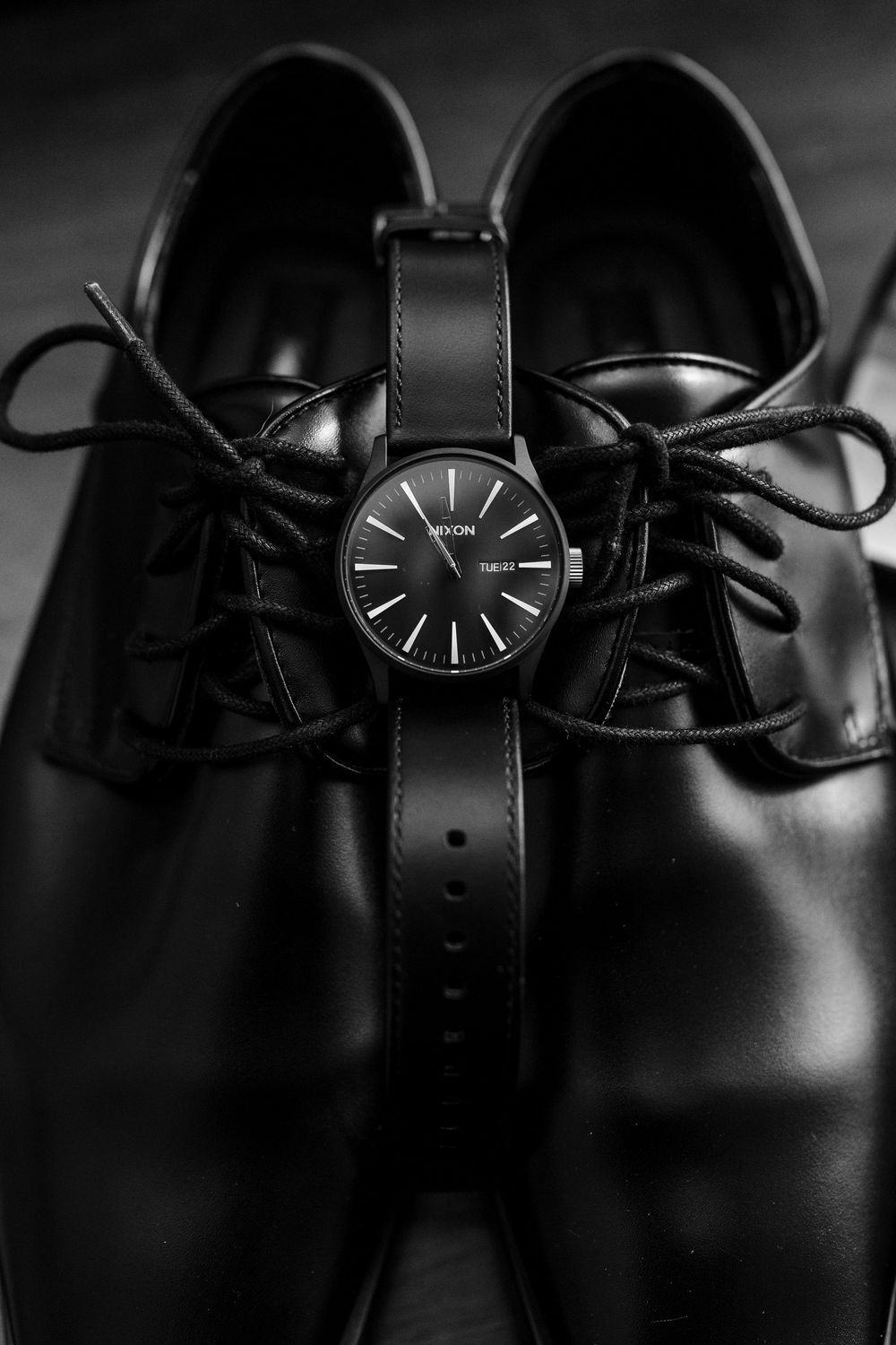 A black wristwatch with minimal design placed on shiny black formal dress shoes against dark background.