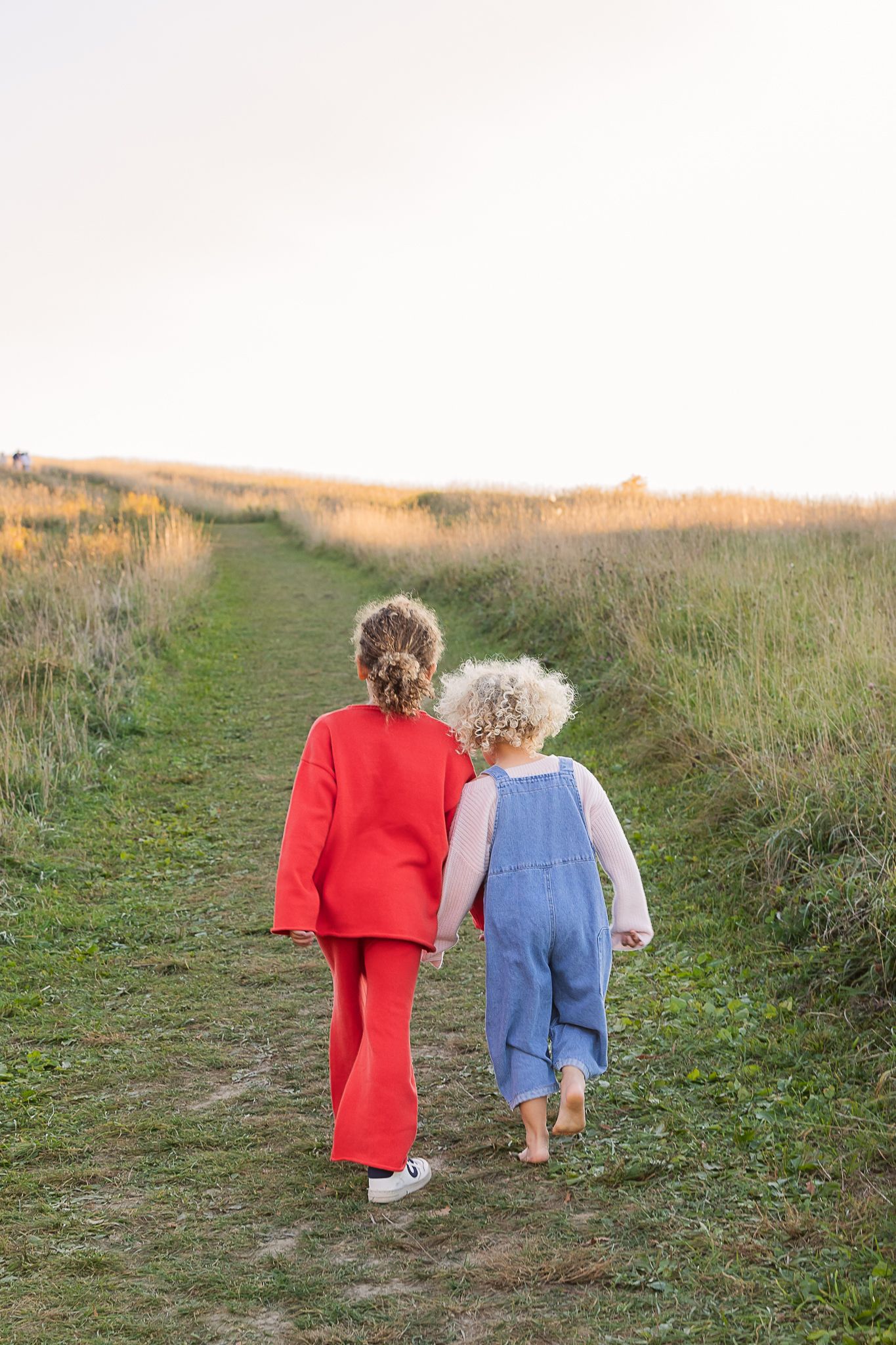 Two children walking down grassy path together, one in red outfit and one in blue overalls.