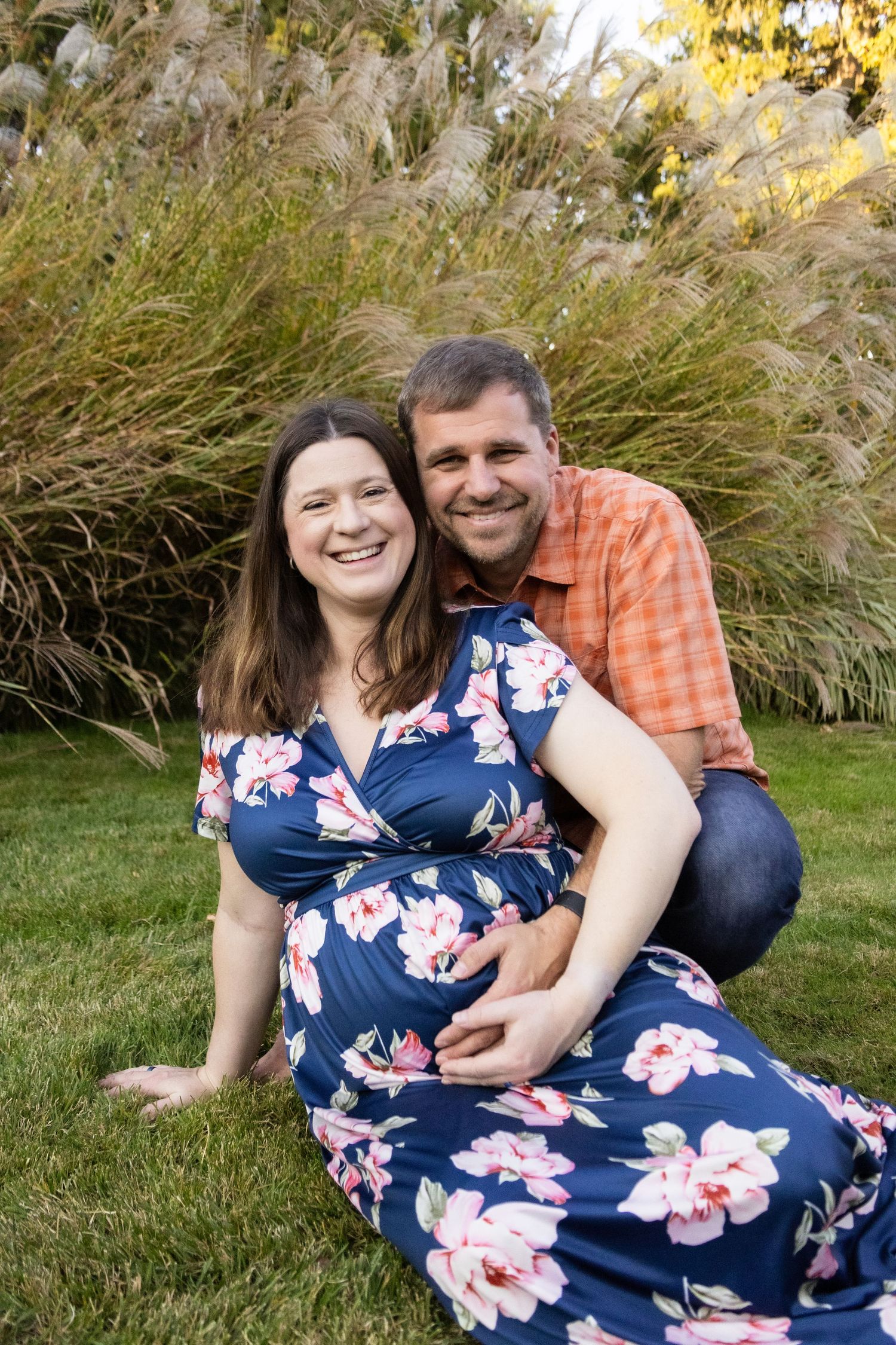 Couple in floral dress and plaid shirt pose in tall grass at sunset.