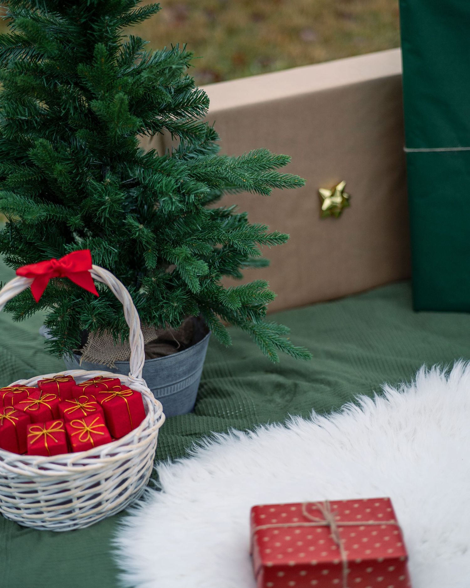 Small Christmas tree with white basket of red ornaments and wrapped present on white furry rug.