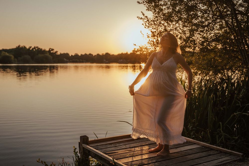 Pregnant woman in white dress standing on wooden dock by lake at golden sunset.