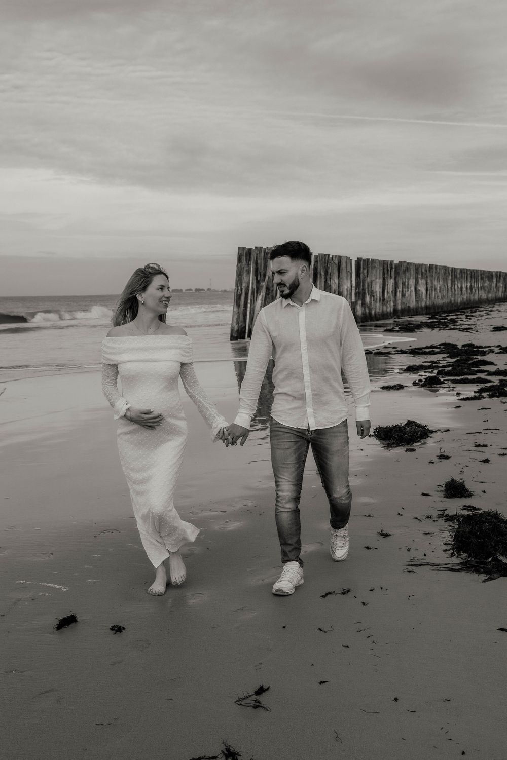 A couple walks hand in hand along a moody black and white beach with wooden posts in the background.