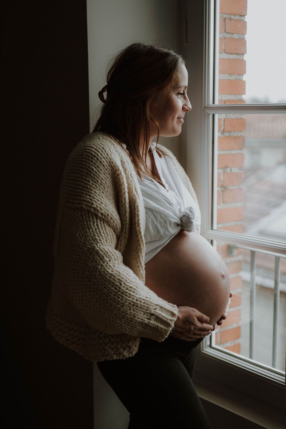 A pregnant person in a beige cardigan sits by a window in a contemplative series of maternity photos.