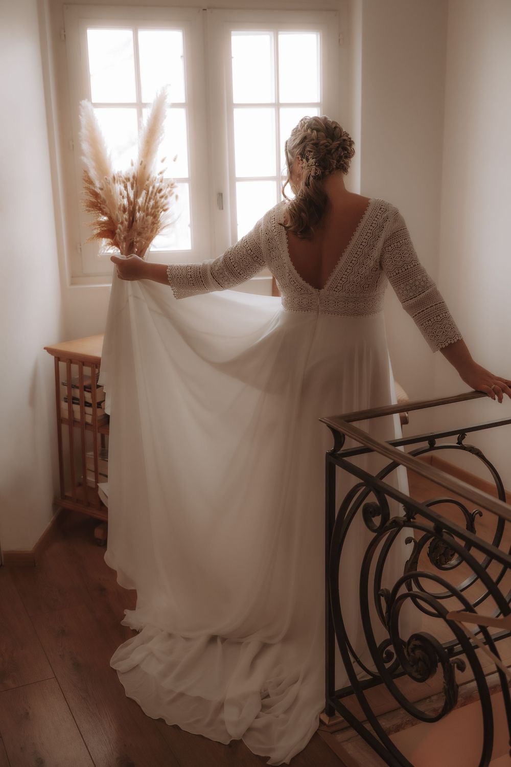 A wedding dress with long lace sleeves and flowing skirt is photographed in soft light by a staircase with dried pampas grass.