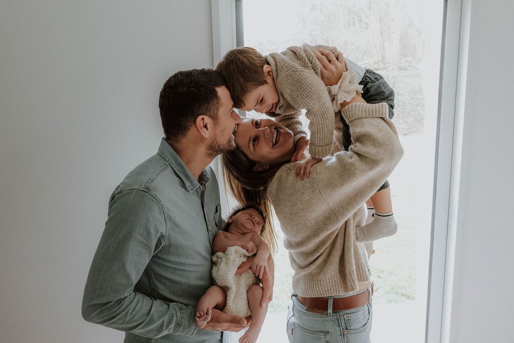 Happy family with father holding newborn baby while mother lifts toddler, sharing kisses indoors.