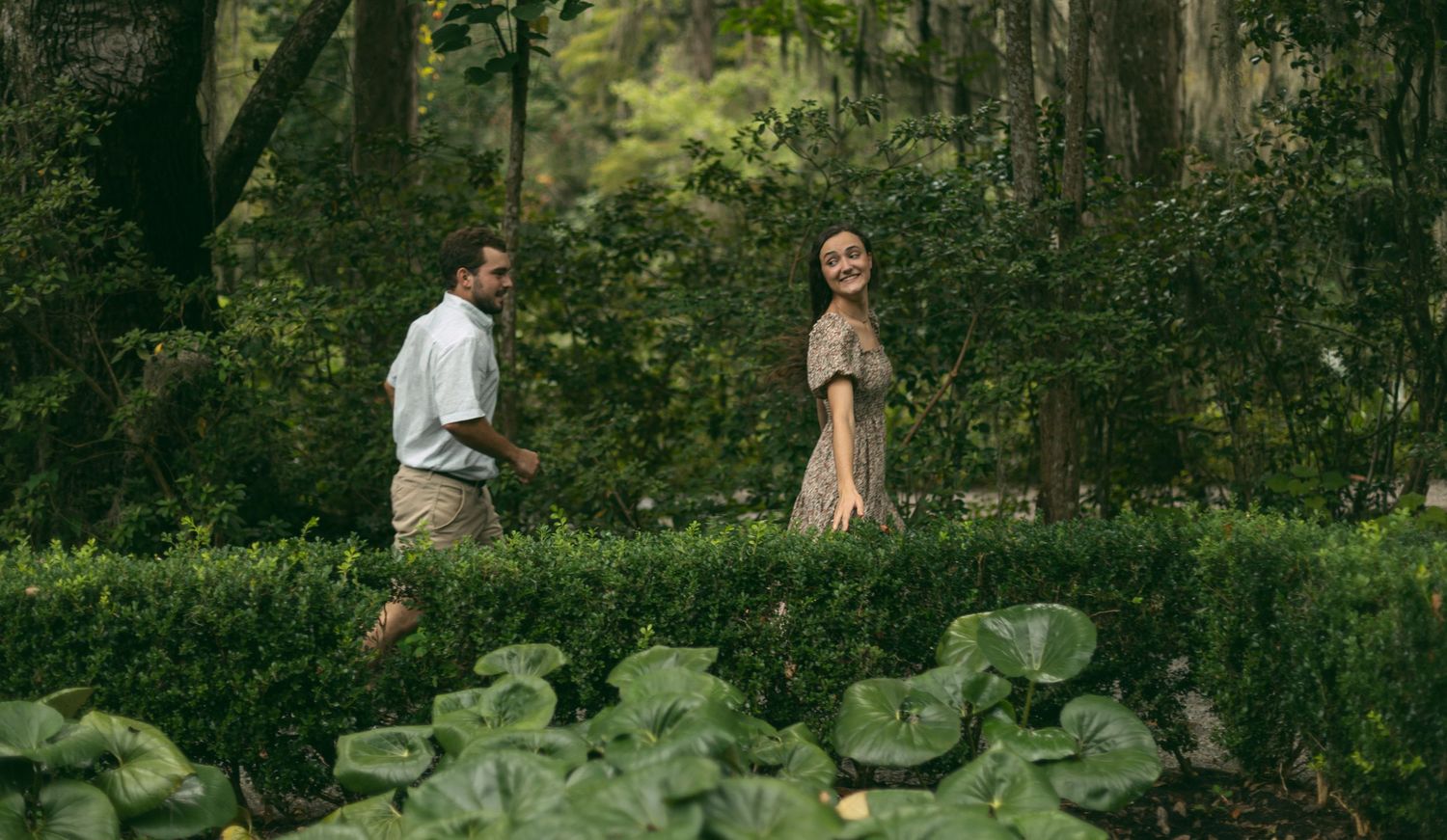 A couple stands together on a garden path surrounded by lush greenery and hedges.