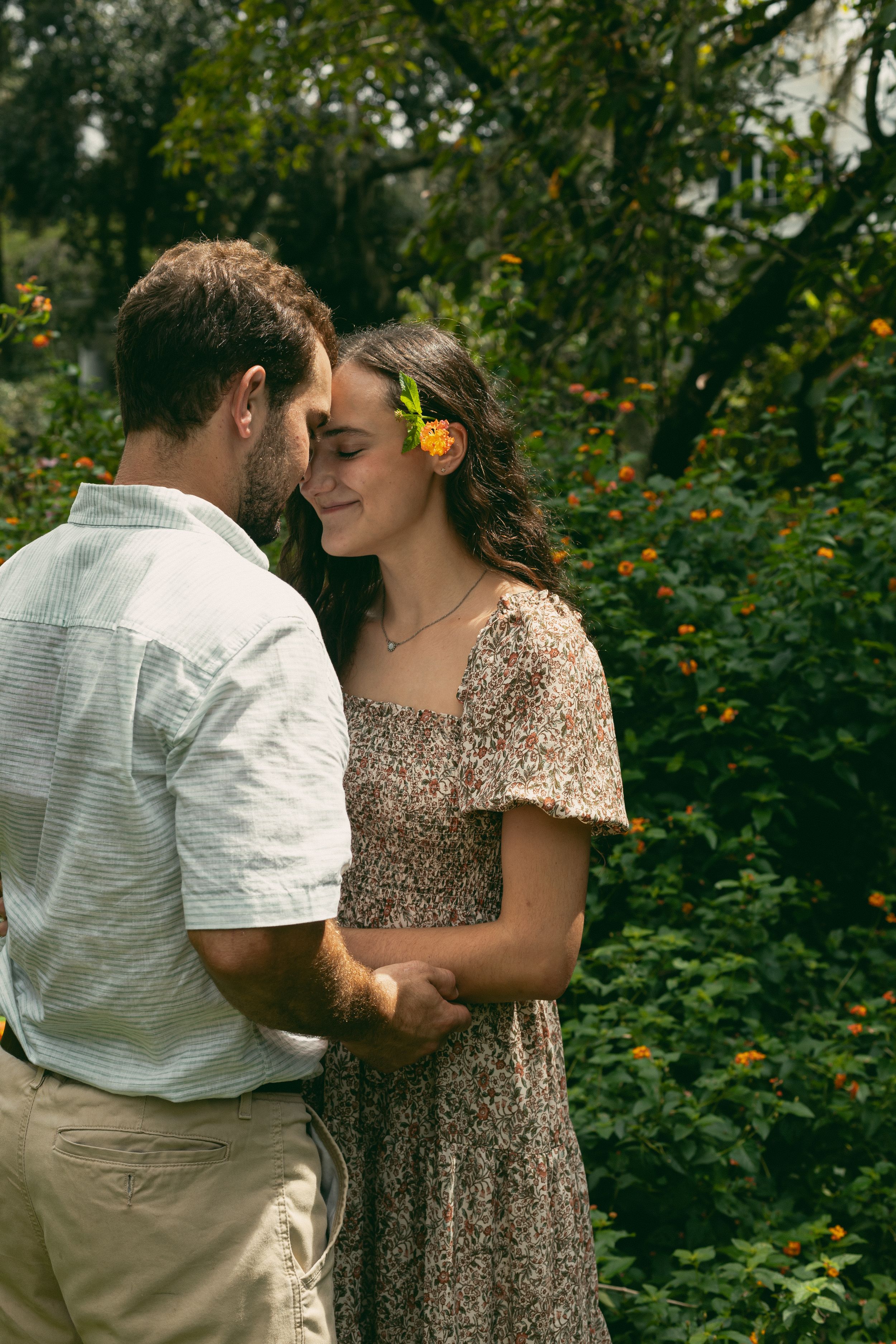 A romantic couple shares an intimate moment in a garden setting with the woman wearing a floral dress.