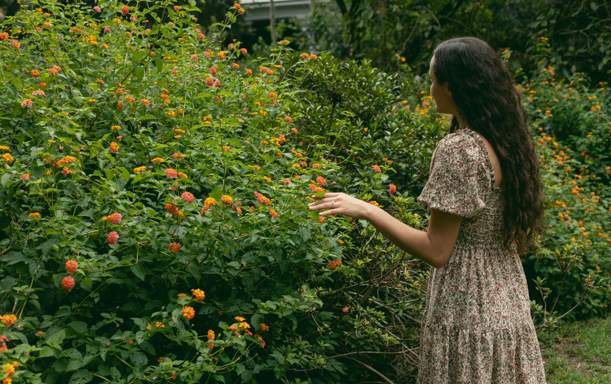 Person in a floral dress admiring yellow flowers in a lush garden setting.
