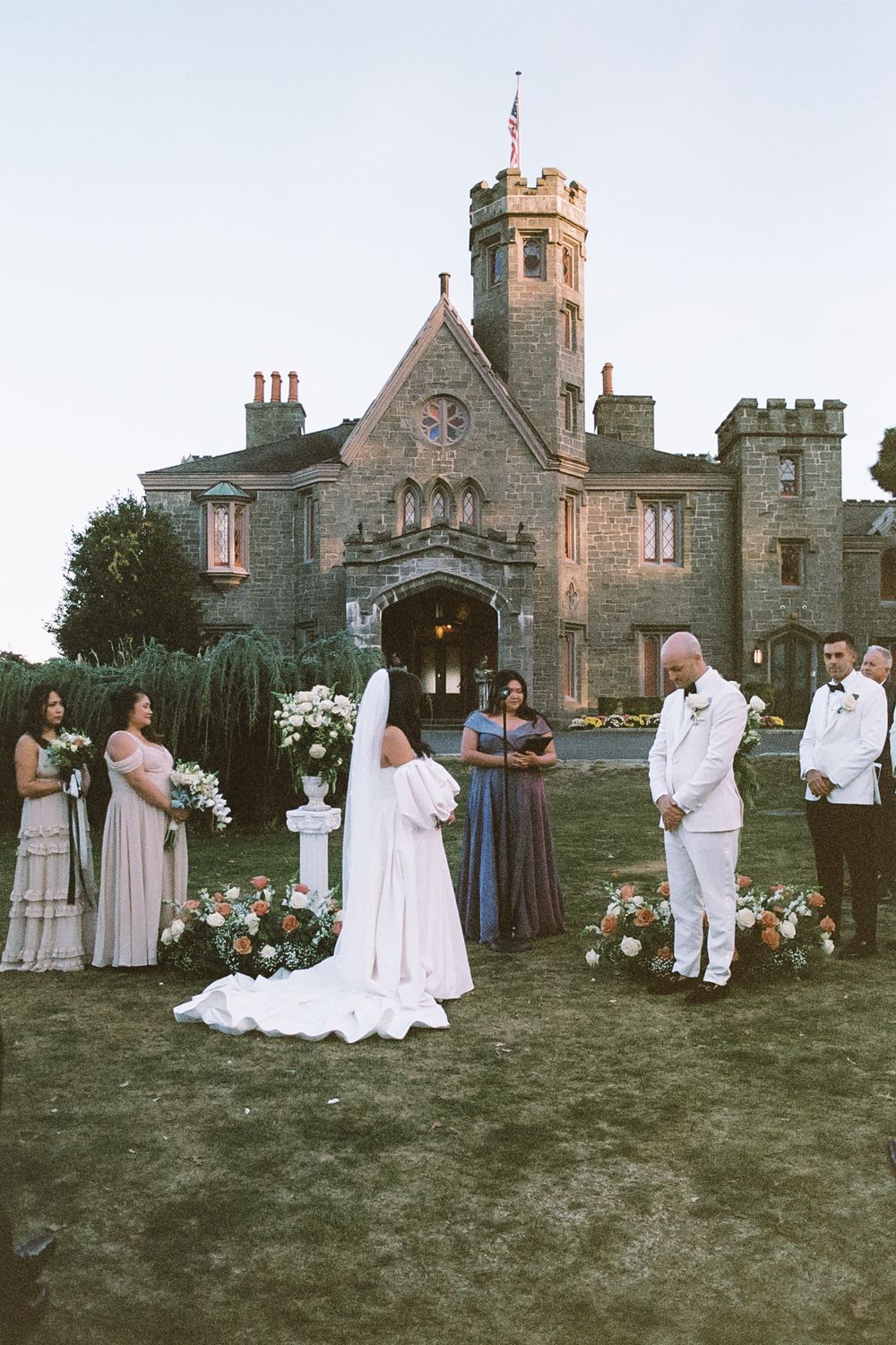 Wedding group in white and pink attire gathered on lawn with gothic architecture backdrop.
