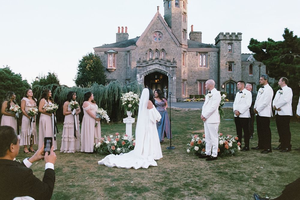 Wedding party poses on lawn in front of gothic stone mansion at sunset.