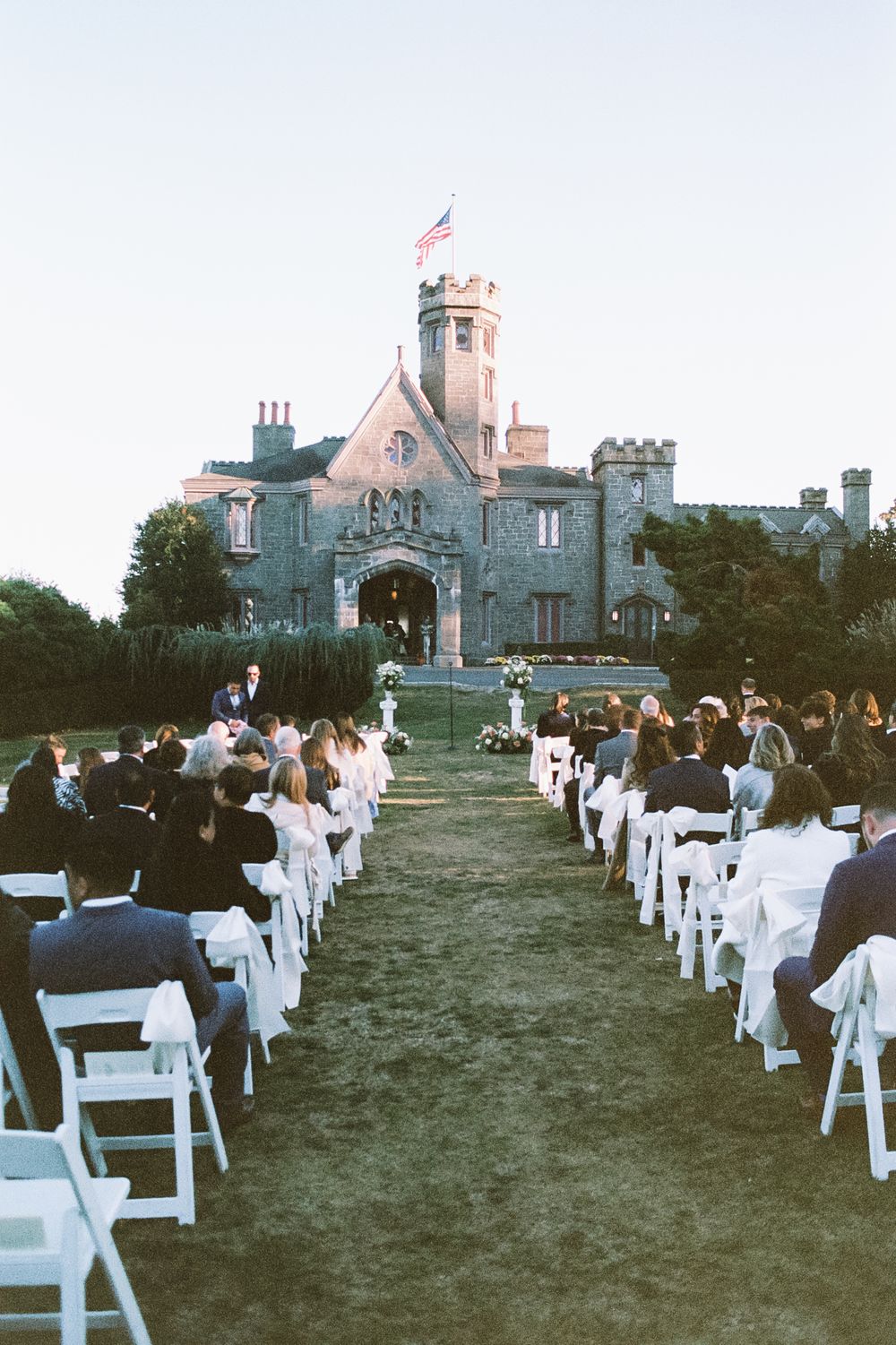 Outdoor wedding ceremony seating arrangement with white chairs facing historic stone building.