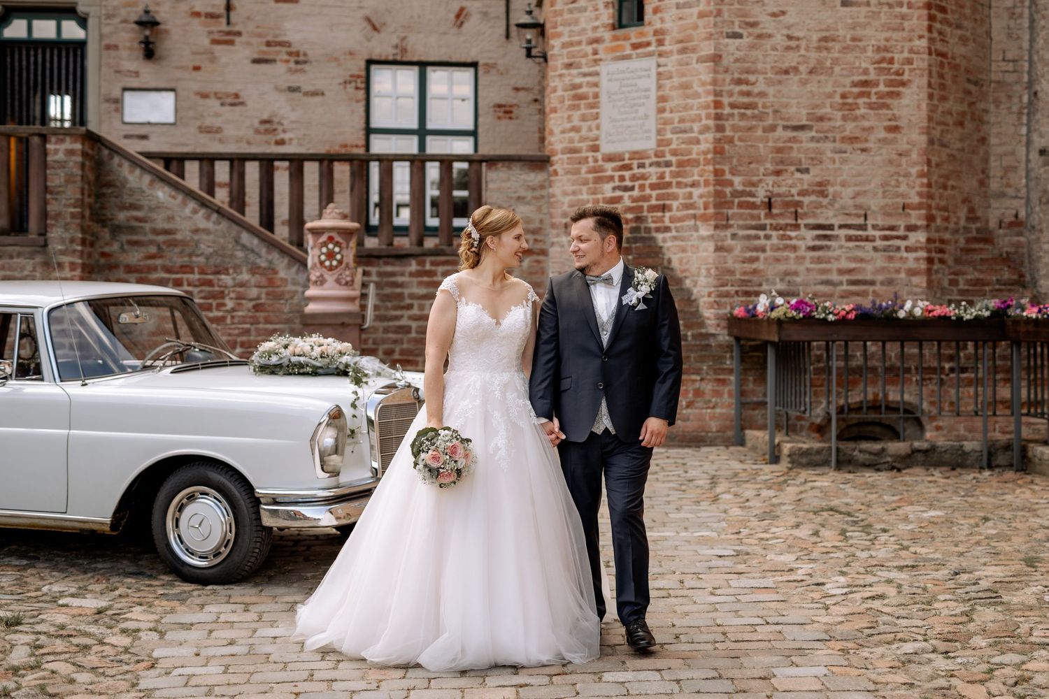 A couple walking hand in hand next to a classic car outside a rustic brick building.