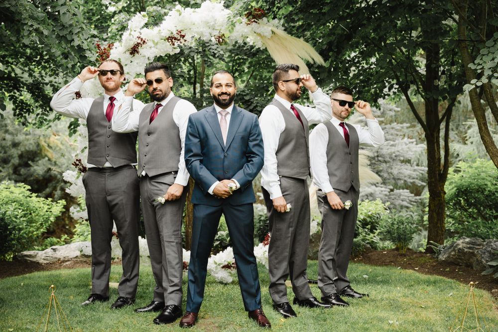 Groomsmen in grey vests stand with the groom in navy blue suit under garden trees.
